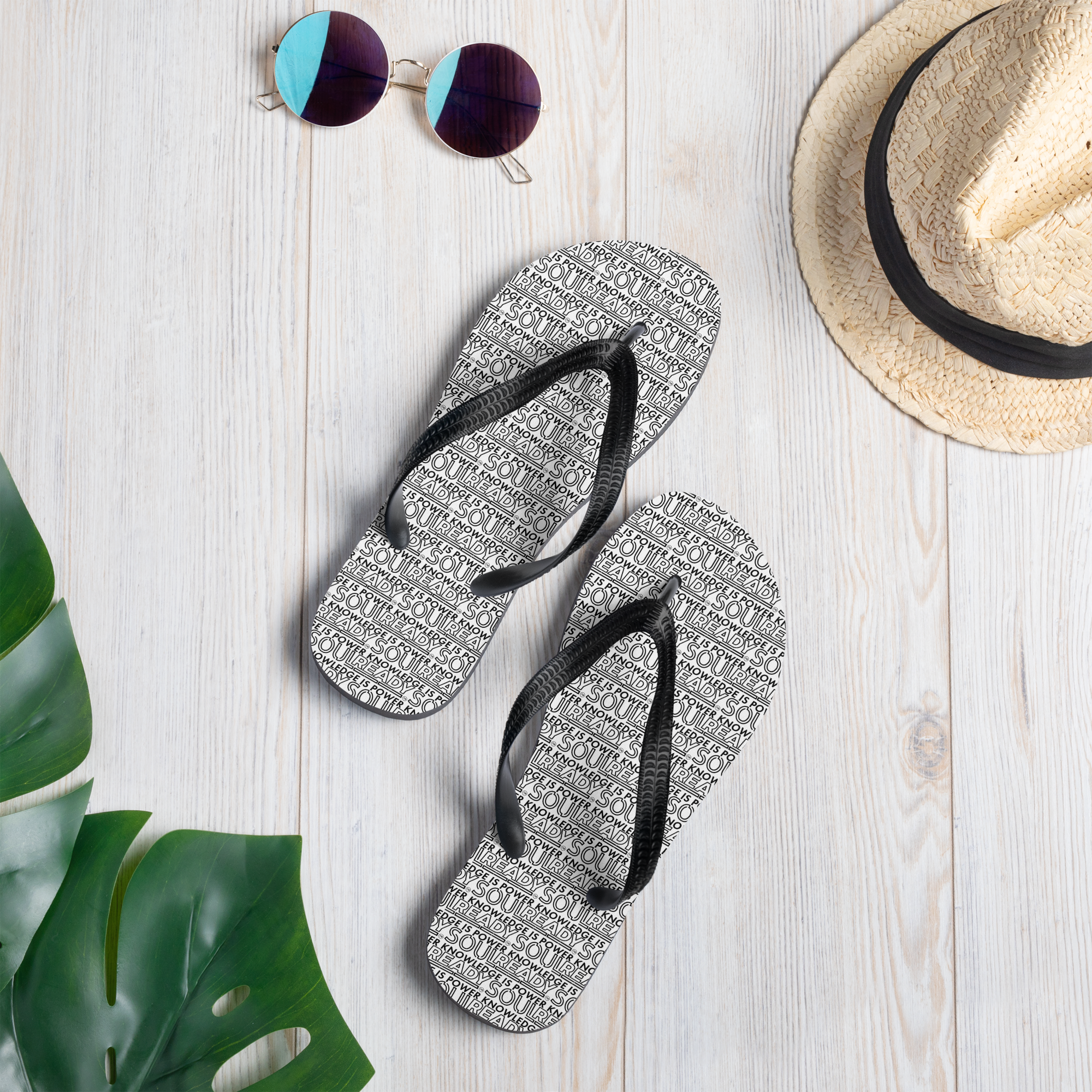 White sublimation flip flops with black straps on wood floor, sunglasses, hat, green leaf nearby